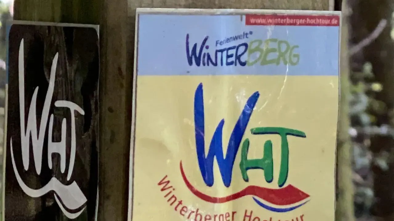 Schilder für die Winterberger Hochtour. Ein Schild zeigt "WHT" in Weiß auf Schwarz, das andere "Winterberg" und "Winterberger Hochtour" in bunten Farben.