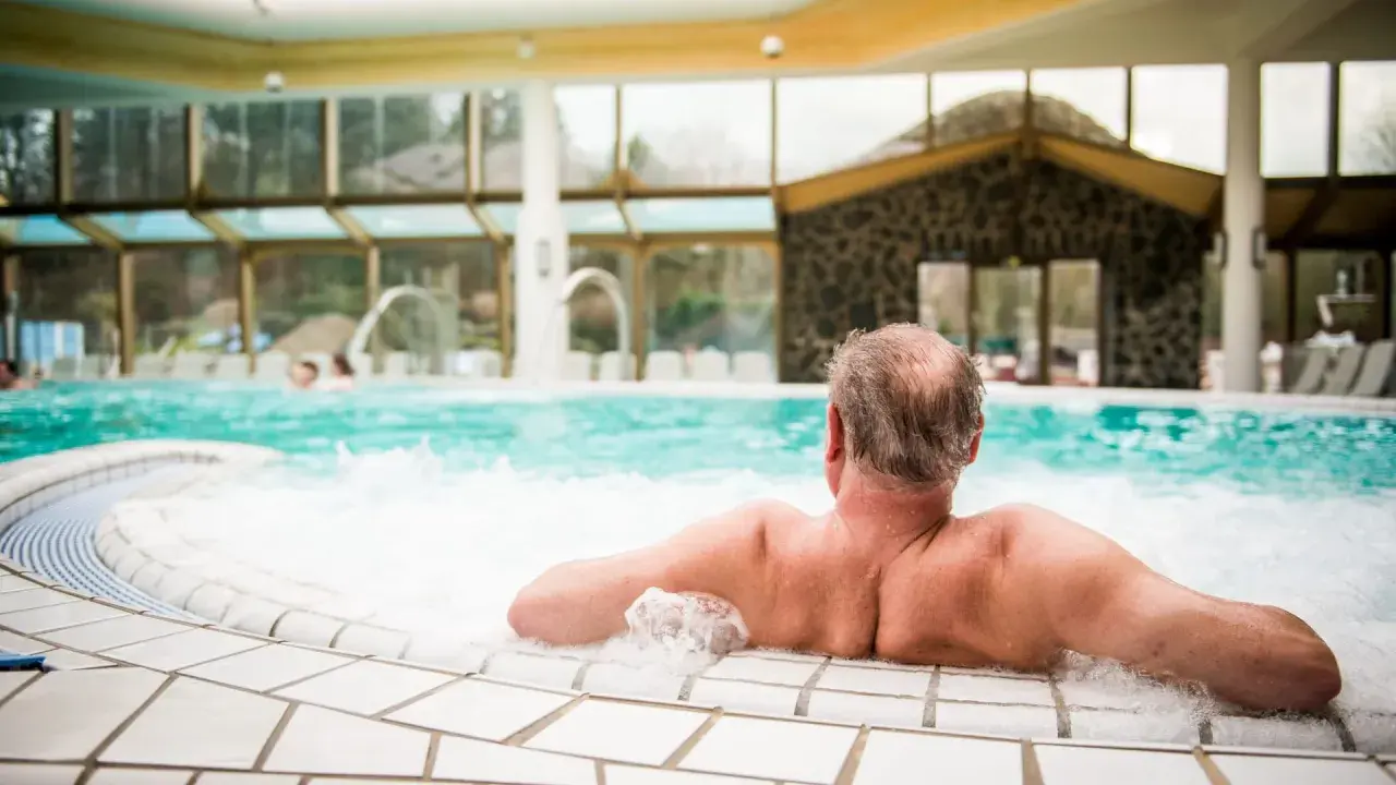 Entspannter Mann genießt das sprudelnde Wasser im mattlerbusch schwimmbad. Hinter ihm ein großes Becken mit Wasserspielen und viel Glas.