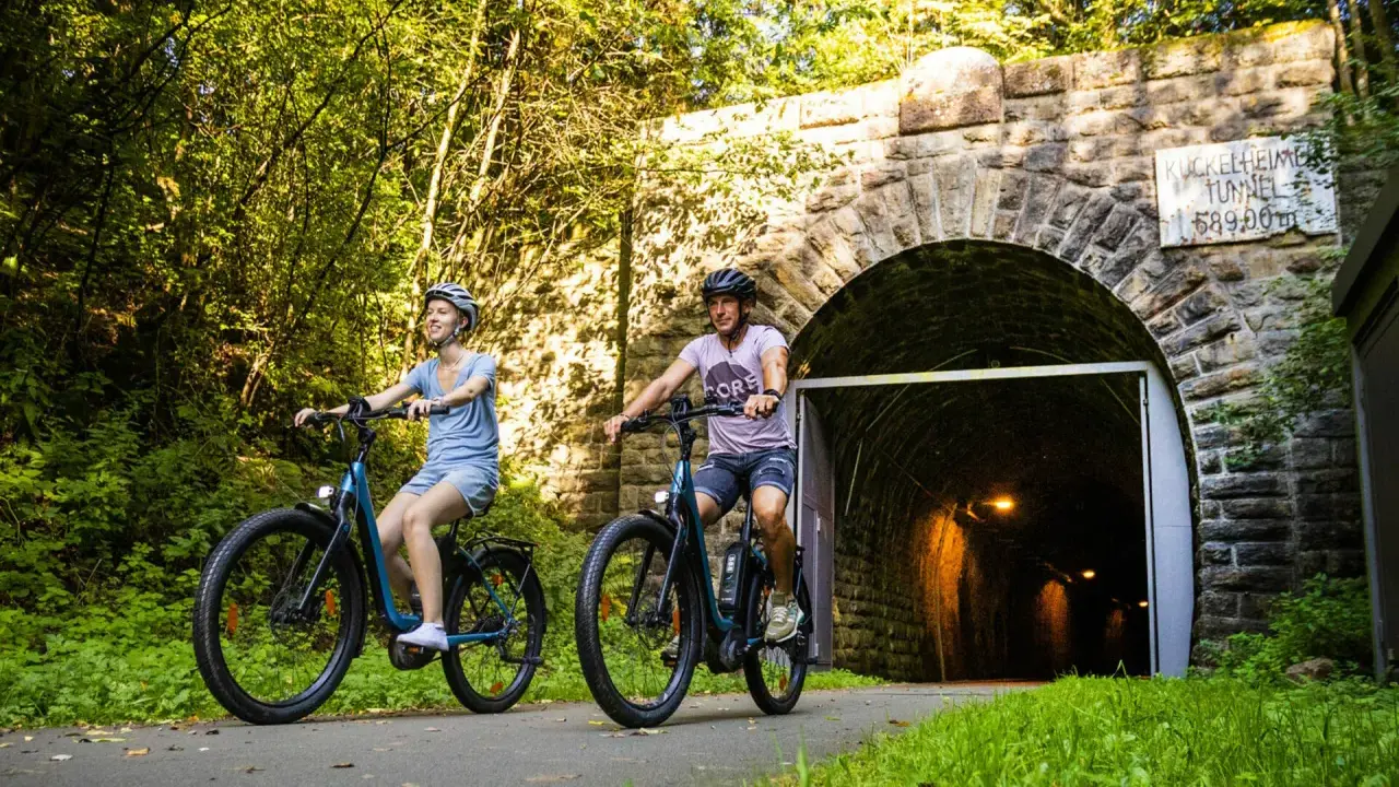 Ein Paar genießt eine entspannte Sauerland Fahrradtour, radelt auf E-Bikes am Eingang des Kuckelheimer Tunnels entlang.