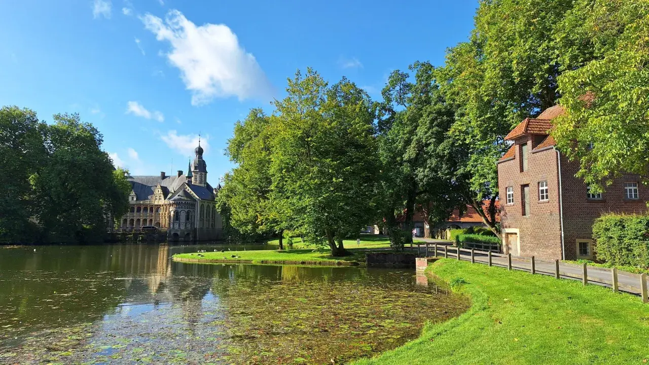 Ein idyllischer vechte radweg führt vorbei an einem Teich, grünen Bäumen und einem historischen Wasserschloss.