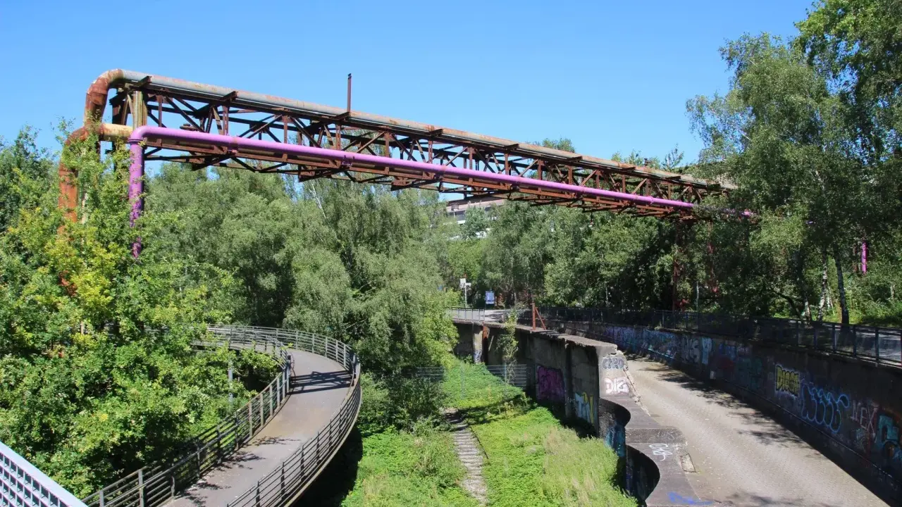Alte Rohrleitungen der Erzbahntrasse Bochum ragen über üppiges Grün. Ein geschwungener Fußweg schlängelt sich durch die Natur.