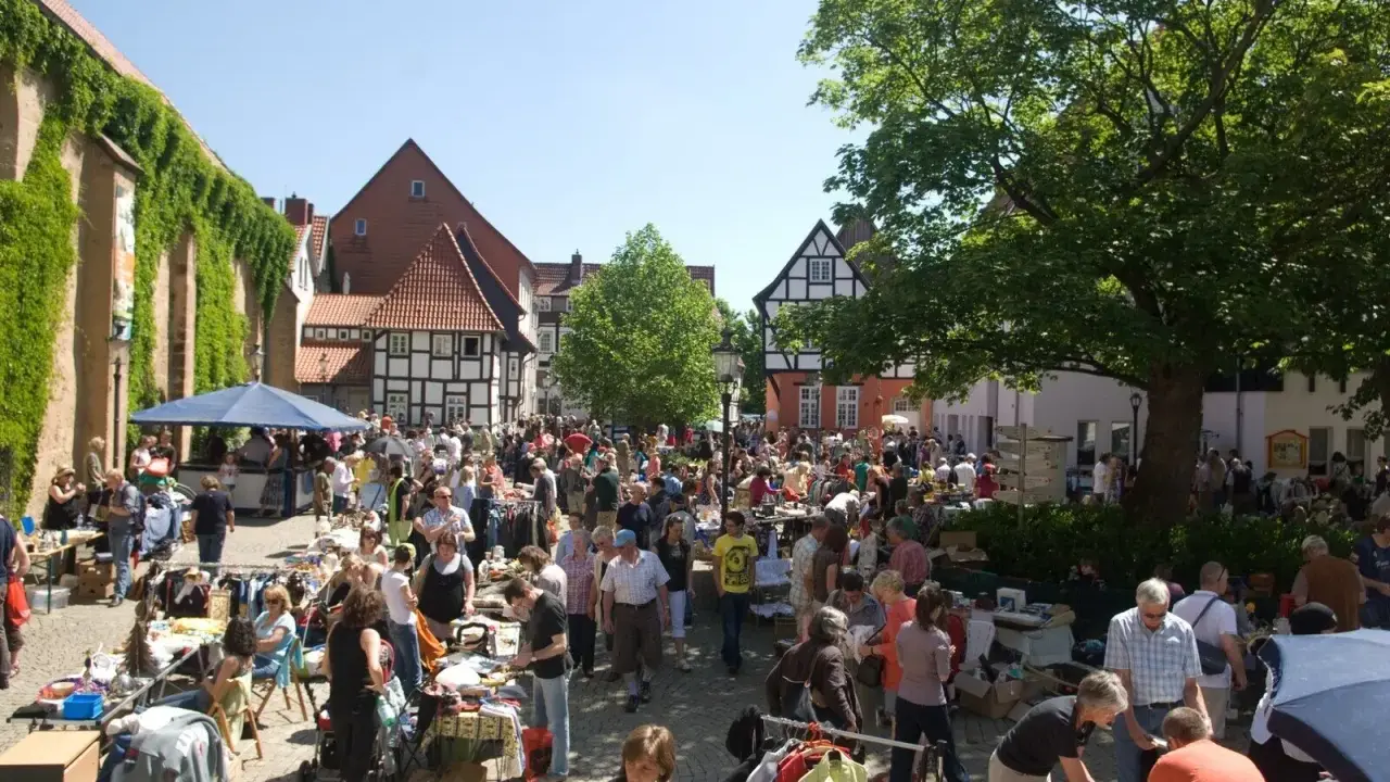 Buntes Treiben auf dem Flohmarkt Minden. Menschen stöbern an Ständen mit Kleidung und Trödel vor Fachwerkhäusern.