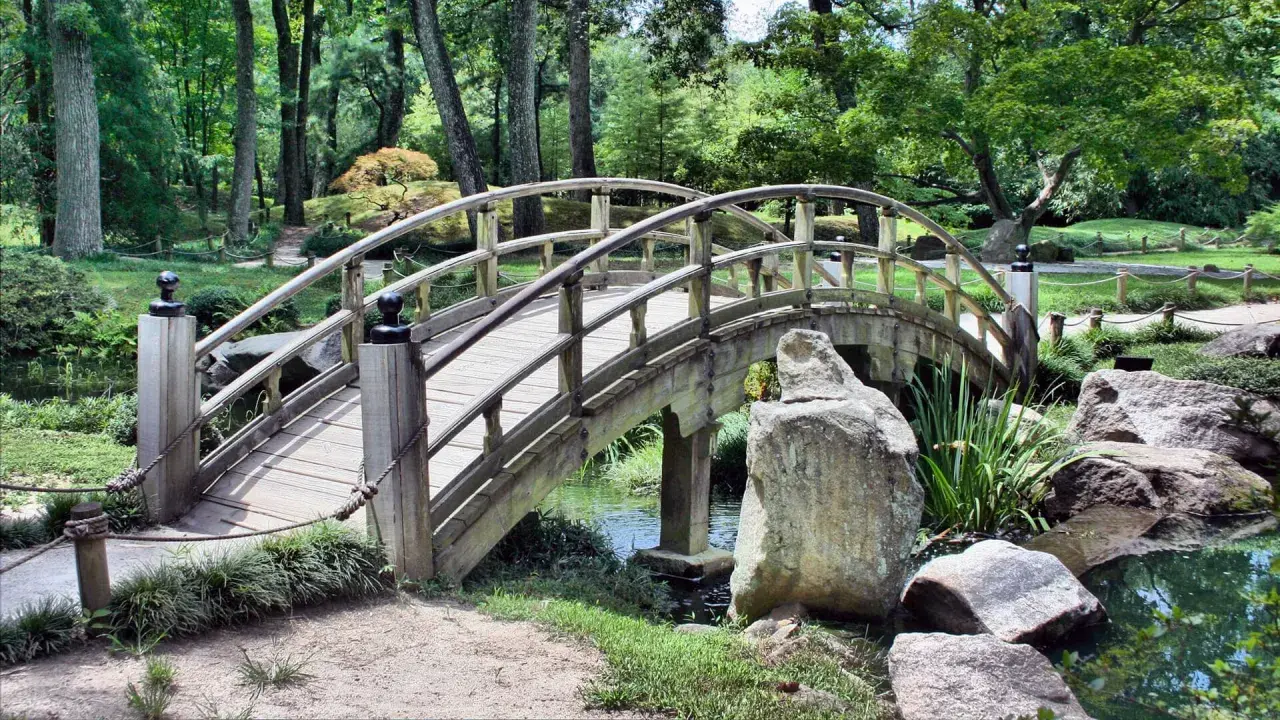 Eine hölzerne Bogenbrücke führt durch den grünen Herford Park. Ein kleiner Bach plätschert unter der Brücke hindurch.