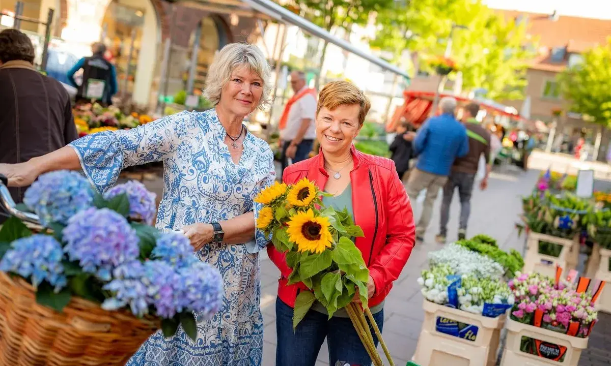 Zwei Frauen mit Blumen auf dem Markt in Coesfeld. Eine hält Sonnenblumen, die andere hat Hortensien im Fahrradkorb.