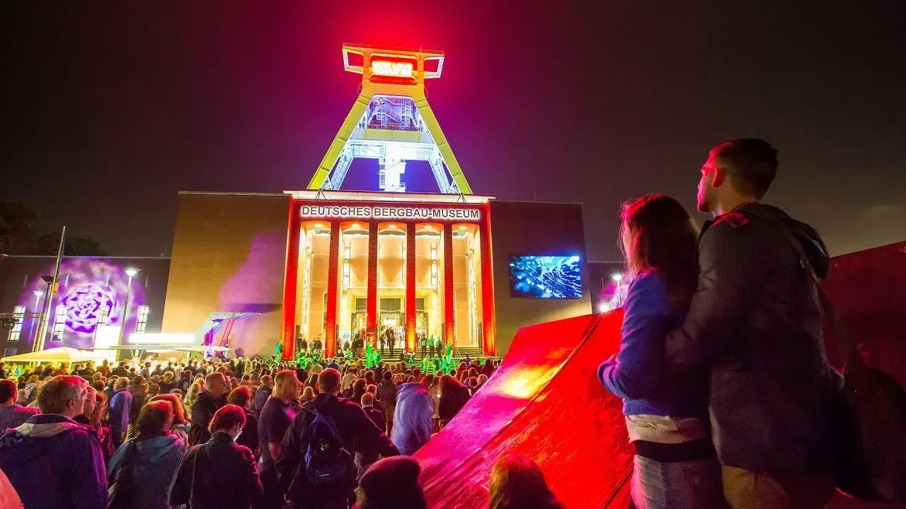 Ein Paar genießt die **Extraschicht Bochum** vor dem beleuchteten Deutschen Bergbau-Museum. Eine Menschenmenge feiert unter dem Förderturm.