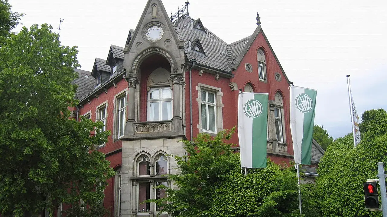 Die Kaiservilla in Minden, ein rotes Backsteingebäude mit Türmchen und zwei GWD-Fahnen, umgeben von üppigem Grün.