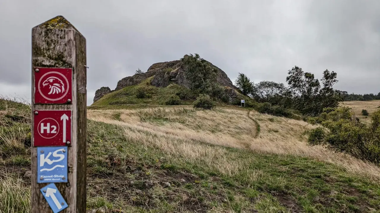 Wanderwegweiser mit Falken-Symbol und "H2"-Markierung, der zu den **rundwanderwege owl** führt. Ein Pfad schlängelt sich über einen grasbewachsenen Hügel zu einer Felsformation.