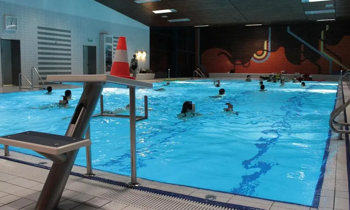 Menschen schwimmen im **Gartenhallenbad Enger**. Ein Sprungbrett mit einem orange-weißen Kegel steht am Beckenrand.