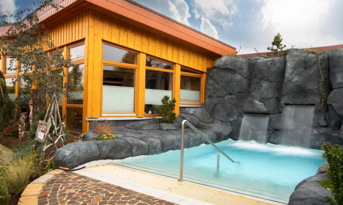 Entspannendes Wasserbecken mit Wasserfällen vor einer Holzhütte, ideal für eine H2O Sauna.