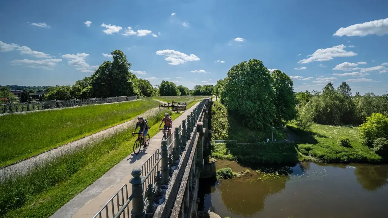Zwei Radfahrer genießen die Aussicht auf der hohen Mark Radroute über eine Brücke, umgeben von grünen Wiesen und Bäumen.
