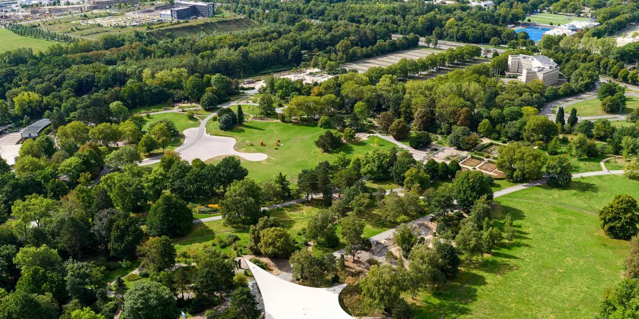 Luftaufnahme eines weitläufigen **Parks in Dortmund** mit grünen Rasenflächen, verschlungenen Wegen, blühenden Beeten und dichtem Baumbestand.