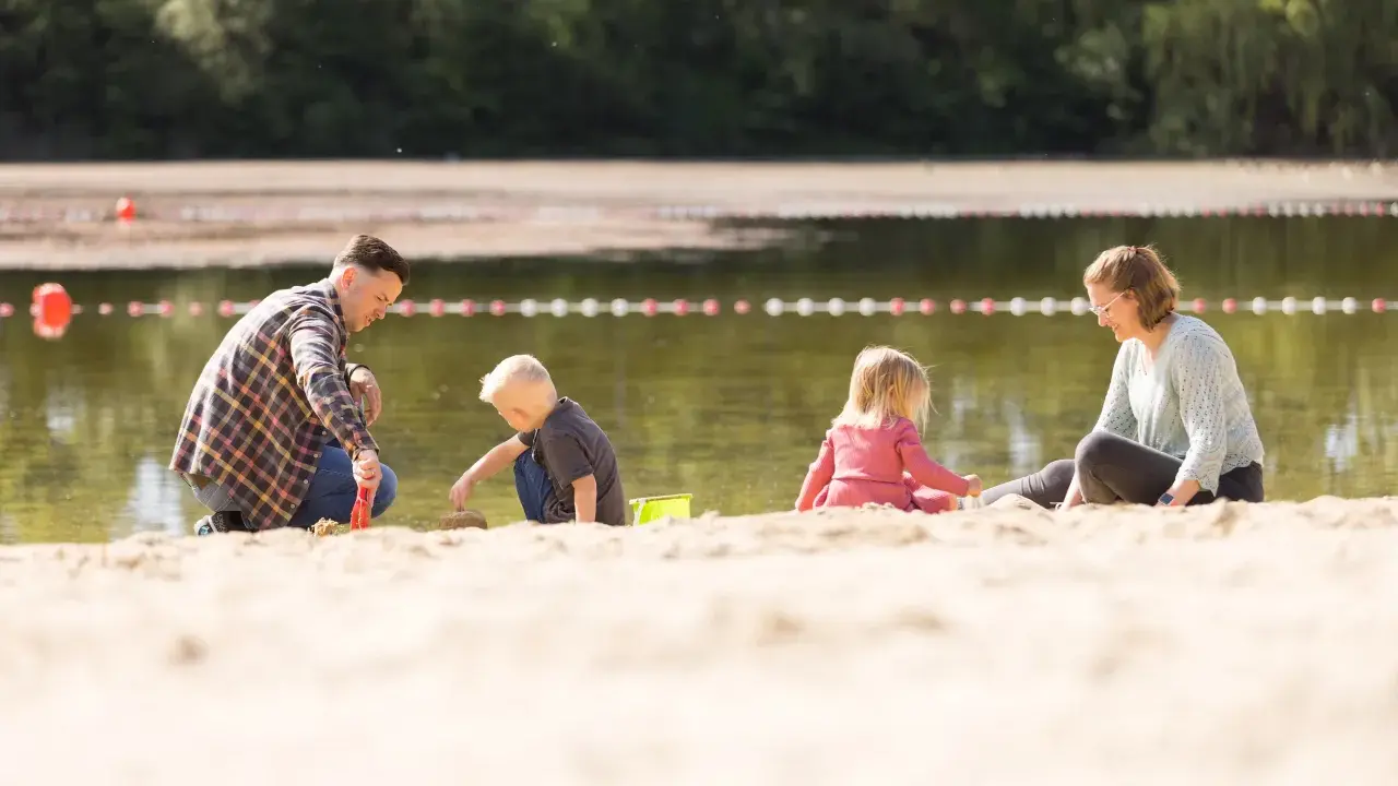 Familie spielt am Sandstrand des badesee mindenerwald. Vater hilft Sohn beim Sandburgen bauen, während Mutter und Tochter daneben sitzen.