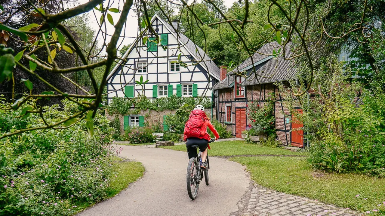Radfahrerin auf einem Weg zwischen Fachwerkhäusern in NRW. Grüne Fensterläden und üppige Vegetation schmücken die idyllische Szenerie.