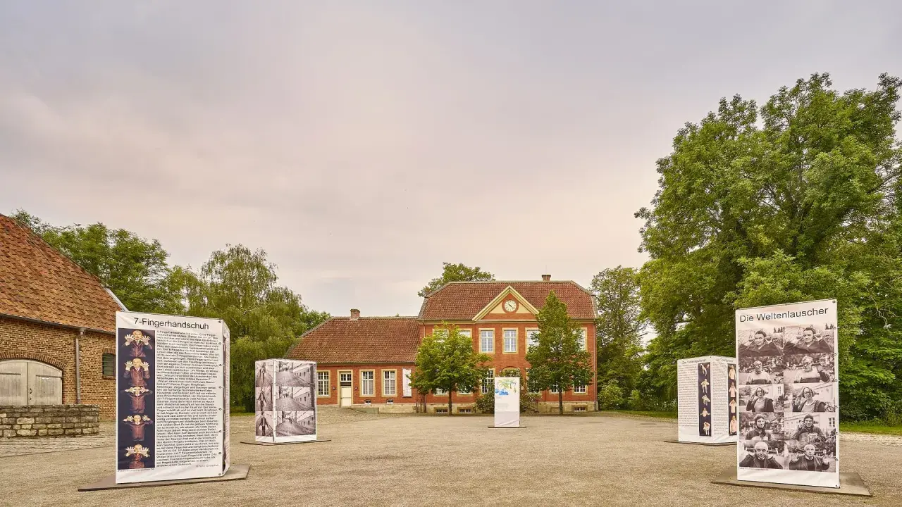 Das Haus Nottbeck mit Ausstellungen im Freien. Mehrere Schilder mit Fotos und Texten stehen vor dem roten Backsteingebäude.