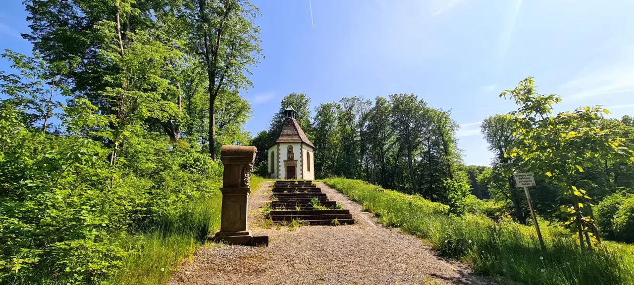 Ein kleiner Weg führt zu einer Kapelle auf einem Hügel, umgeben von grünen Bäumen. Der schmetterlingspfad willebadessen lädt zum Entdecken ein.