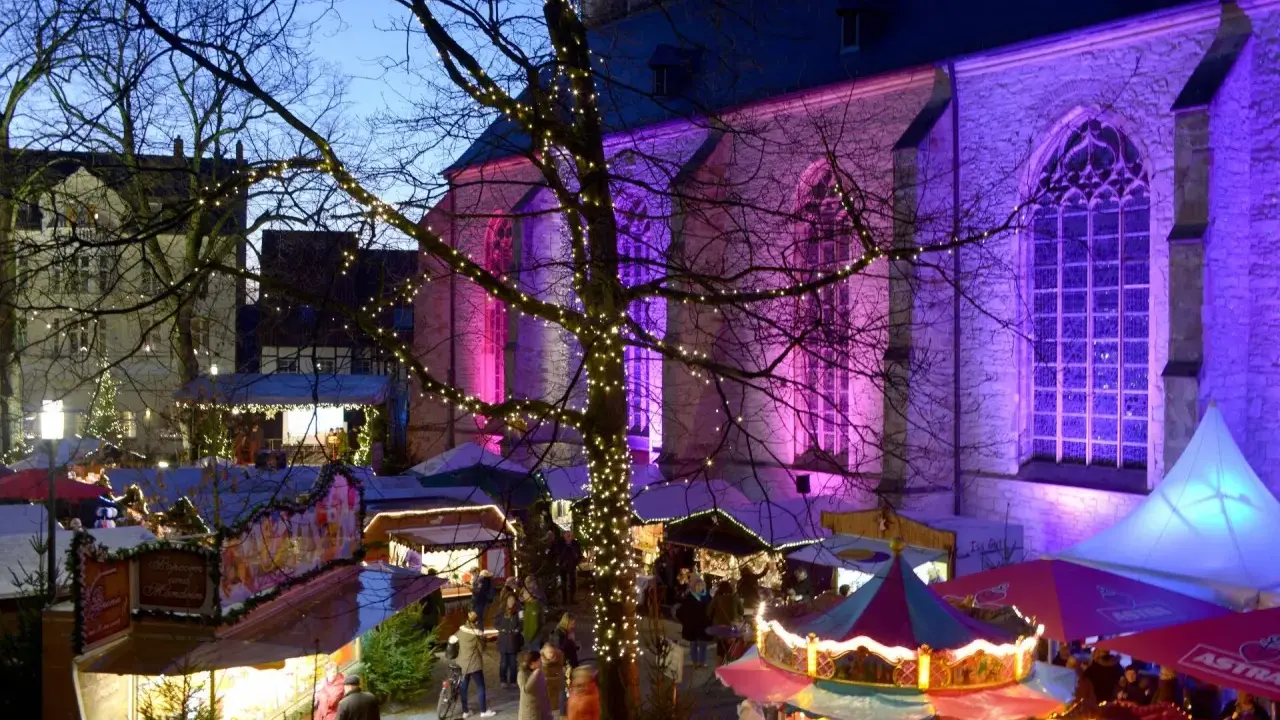 Bunte Lichter und Buden auf dem weihnachtsmarkt werne. Eine Kirche im Hintergrund erstrahlt in lila Licht.