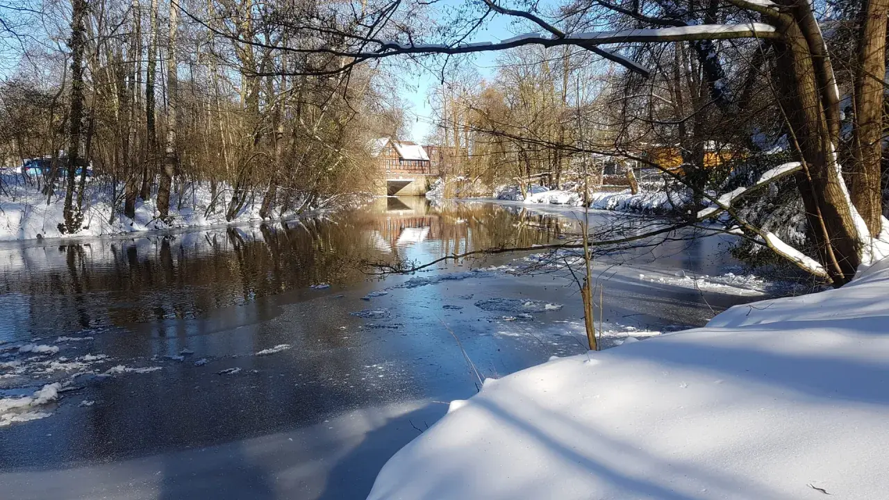 Winterliche Idylle an der **Planwiese Telgte**: Eisiger Fluss, schneebedeckte Ufer und ein Fachwerkhaus spiegeln sich im Wasser.