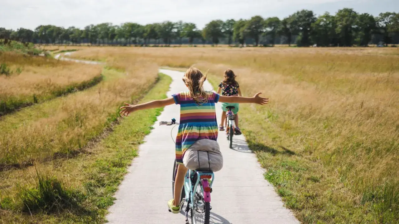 Zwei Mädchen genießen ein sonniges Fahrrad-Wochenende auf einem Feldweg, die Arme weit ausgebreitet.