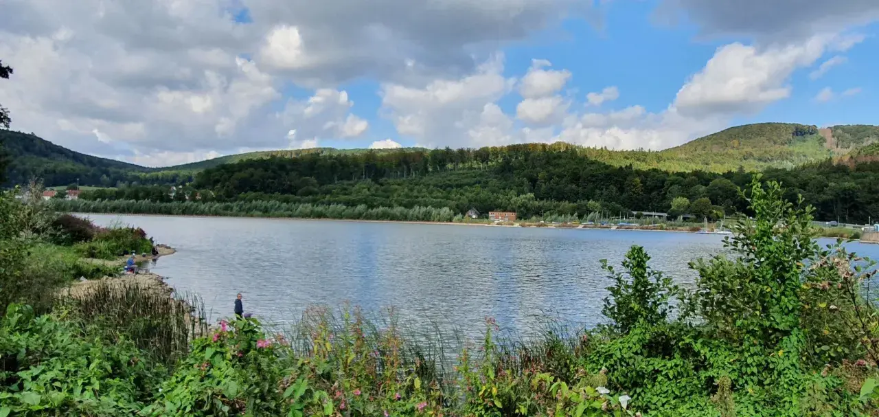 Idyllische Landschaft am Emmer-Radweg mit See, bewaldeten Hügeln und einigen Anglern am Ufer.
