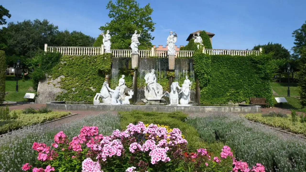 Prachtvoller Brunnen mit Skulpturen und Wasserfällen, umgeben von blühenden Blumen und Grün im botanischen Garten NRW.