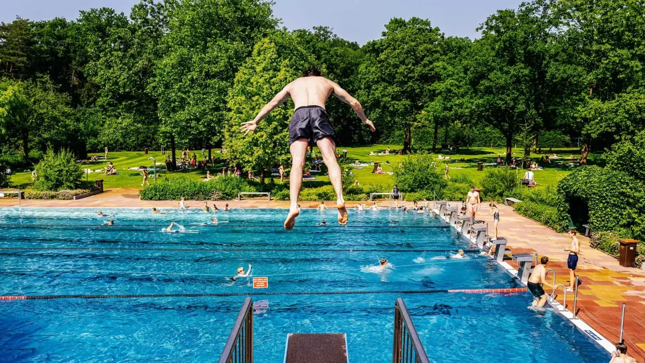 Ein Mann springt in ein Freibad in NRW. Viele Menschen sonnen sich auf der grünen Liegewiese im Hintergrund.