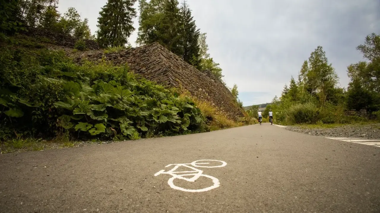 Radweg mit Radsymbol auf Asphalt. Zwei Radfahrer fahren auf einem asphaltierten Weg, der an einer steilen, mit Steinen bedeckten Böschung und üppiger Vegetation vorbeiführt.