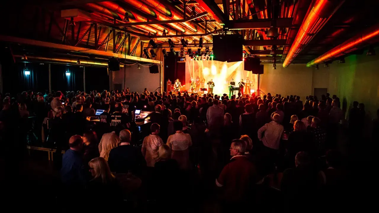 Ein Konzert in Emsdetten mit einer Band auf der Bühne und vielen begeisterten Zuschauern. Die Beleuchtung taucht die Szene in ein warmes, rotes Licht.