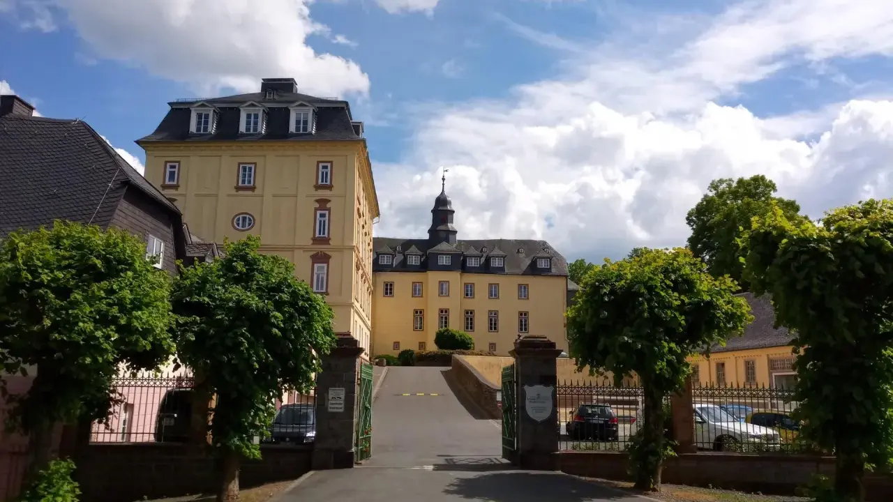 Schloss Wittgenstein in Bad Laasphe, ein gelbes Barockschloss mit Turm, umgeben von Bäumen und einem blauen Himmel mit Wolken.