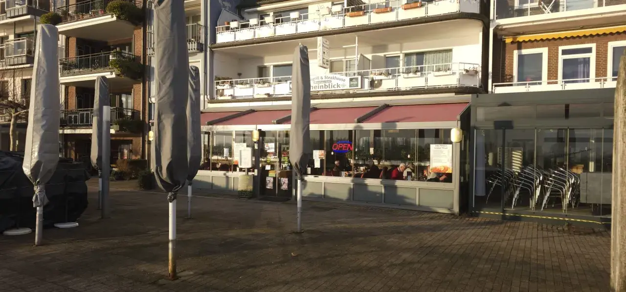 Geschlossene Sonnenschirme säumen die rheinpromenade emmerich. Ein Restaurant mit "OPEN"-Schild lädt zum Verweilen ein.