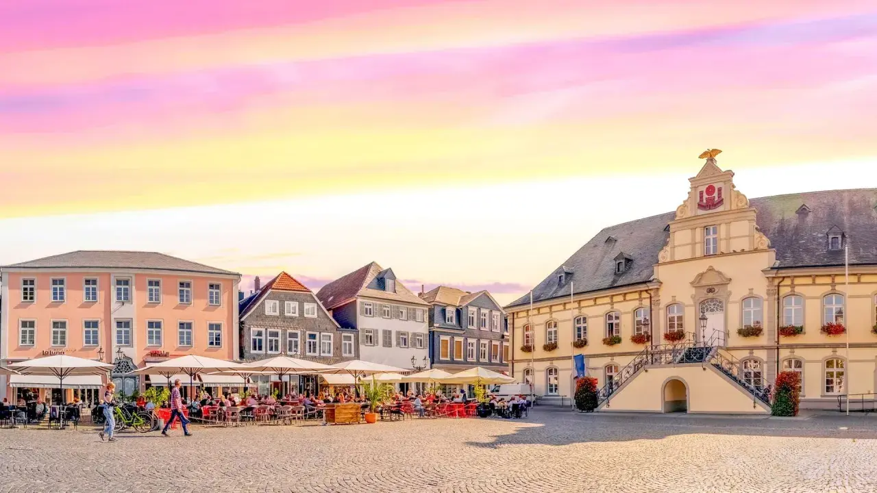Ein Marktplatz mit Cafés und historischen Gebäuden, der an das "Venedig Westfalens" erinnert, unter einem farbenfrohen Himmel.