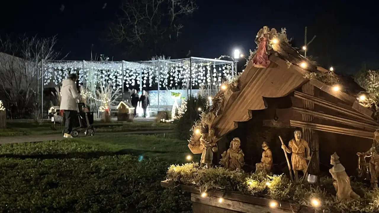 Bethlehemskrippe mit Figuren und Lichtern auf dem weihnachtsmarkt legden. Im Hintergrund eine Person mit Rollator und Lichterketten.