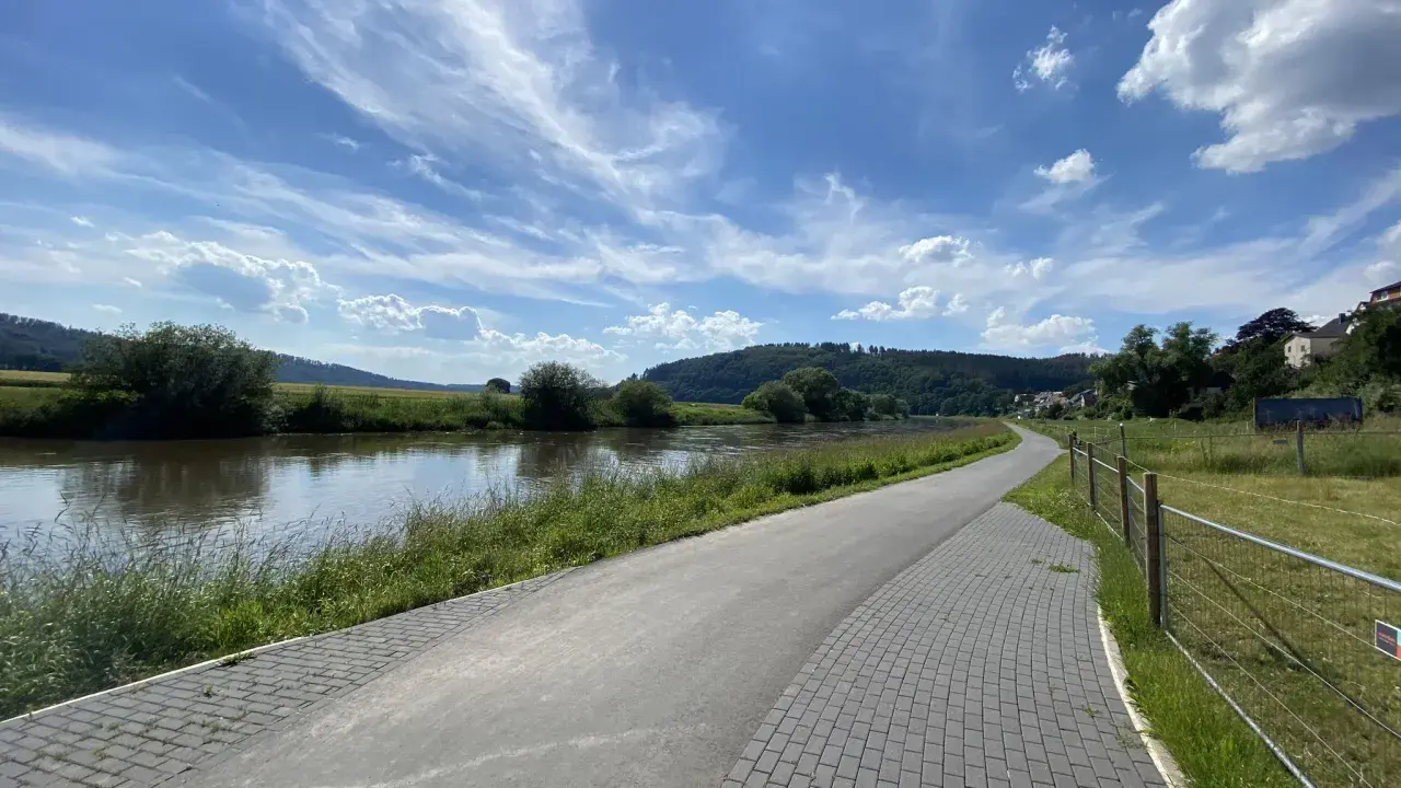 Ein asphaltierter Weg säumt den Fluss, ideal für eine Weser-Radweg Rundtour. Grüne Ufer und ein blauer Himmel mit Wolken laden zum Radfahren ein.