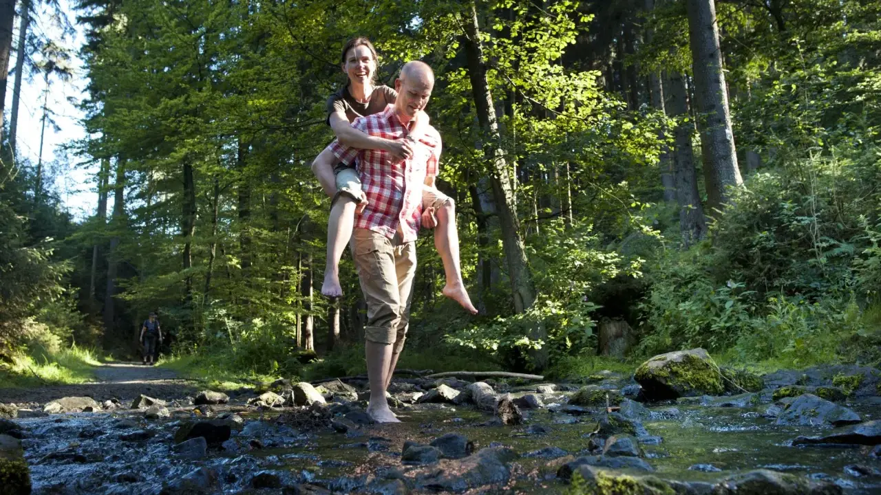 Ein Mann trägt eine Frau auf dem Rücken durch einen Bach im Wald. Die Sonne scheint durch die Bäume. Ein idyllischer Ort, wie eine teuto quelle.