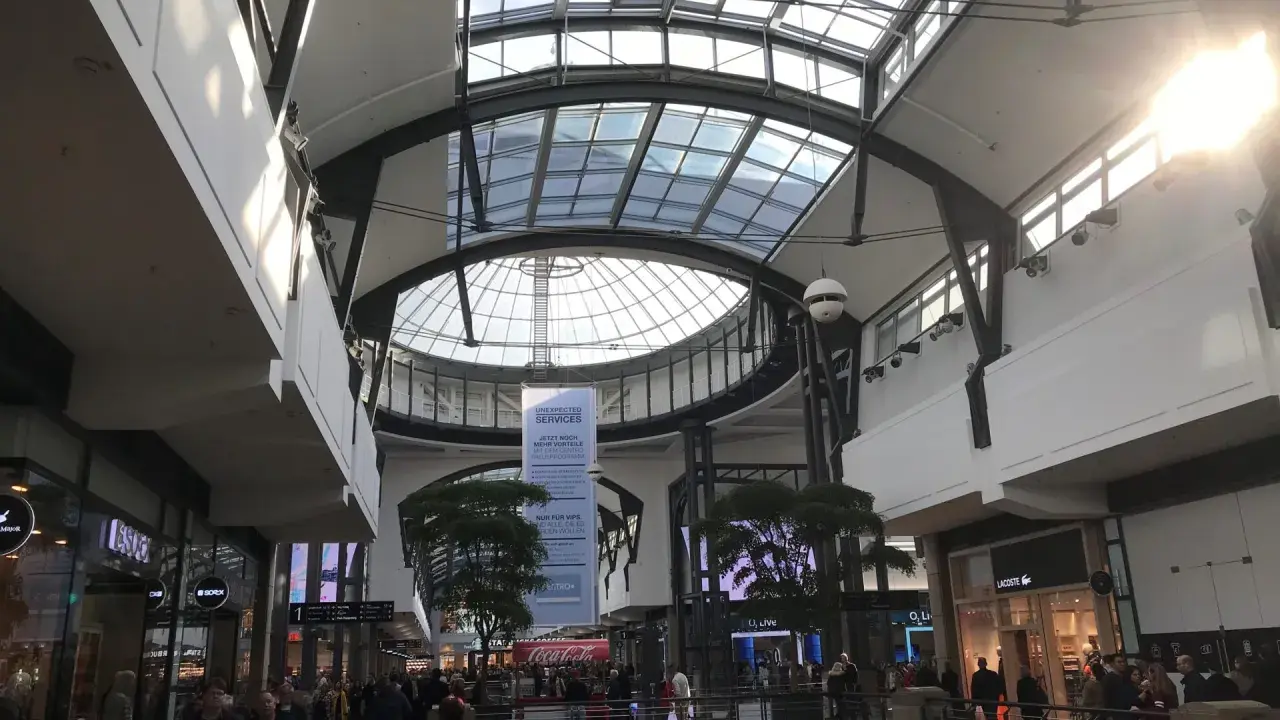 Helles, modernes Einkaufszentrum mit Glasdach. Menschen schlendern durch das Oberhausen Centro, vorbei an Geschäften wie Lacoste.