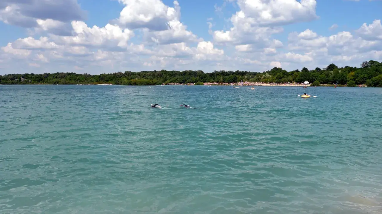 Schwimmer im türkisfarbenen Wasser des Beckum Sees, mit bewaldetem Ufer und Wolken am Himmel.