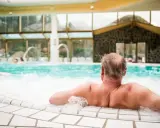 Entspannter Mann genießt das sprudelnde Wasser im mattlerbusch schwimmbad. Hinter ihm ein großes Becken mit Wasserspielen und viel Glas.