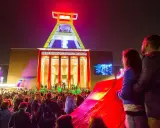 Ein Paar genießt die **Extraschicht Bochum** vor dem beleuchteten Deutschen Bergbau-Museum. Eine Menschenmenge feiert unter dem Förderturm.