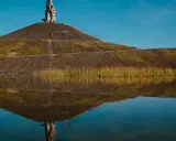 Spiegelung einer Halde im Ruhrgebiet mit einer Steinskulptur auf dem Gipfel und Schilf am Ufer.