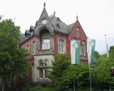 Die Kaiservilla in Minden, ein rotes Backsteingebäude mit Türmchen und zwei GWD-Fahnen, umgeben von üppigem Grün.