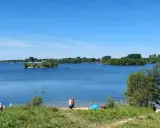 Sonniger Tag am Lippesee Paderborn. Menschen entspannen am Ufer, Segelboote gleiten über das Wasser, eine kleine Insel ziert die Mitte.