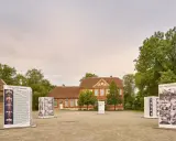 Das Haus Nottbeck mit Ausstellungen im Freien. Mehrere Schilder mit Fotos und Texten stehen vor dem roten Backsteingebäude.