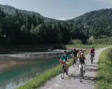Gruppe Radfahrer auf einem Weg entlang eines Flusses mit bewaldeten Hügeln im Hintergrund.