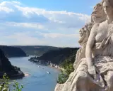 Steinerne Nixe blickt auf den Rhein. Ein malerischer Abschnitt für die Rheinradweg Etappen, mit Schiffen auf dem Wasser und bewaldeten Hängen.