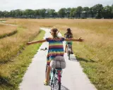 Zwei Mädchen genießen ein sonniges Fahrrad-Wochenende auf einem Feldweg, die Arme weit ausgebreitet.