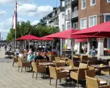 Gemütliche Straßencafés mit roten Sonnenschirmen laden zum Verweilen ein. Menschen spazieren am Rhein entlang, im Hintergrund die Brücke von Emmerich.