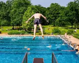 Ein Mann springt in ein Freibad in NRW. Viele Menschen sonnen sich auf der grünen Liegewiese im Hintergrund.