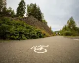 Radweg mit Radsymbol auf Asphalt. Zwei Radfahrer fahren auf einem asphaltierten Weg, der an einer steilen, mit Steinen bedeckten Böschung und üppiger Vegetation vorbeiführt.