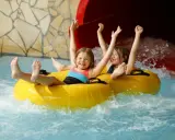 Zwei lachende Kinder rutschen auf einem gelben Schlauchboot durch einen Wasserpark. Ein perfekter Tag, wie ein h2o gutschein!