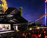 Ein belebtes **freiluft kino** mit vielen Zuschauern, die einen Film auf einer großen Leinwand vor einer beeindruckenden Industriekulisse mit bunten Türmen und Stahlkonstruktionen genießen.