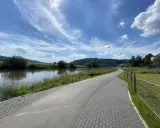 Ein asphaltierter Weg säumt den Fluss, ideal für eine Weser-Radweg Rundtour. Grüne Ufer und ein blauer Himmel mit Wolken laden zum Radfahren ein.